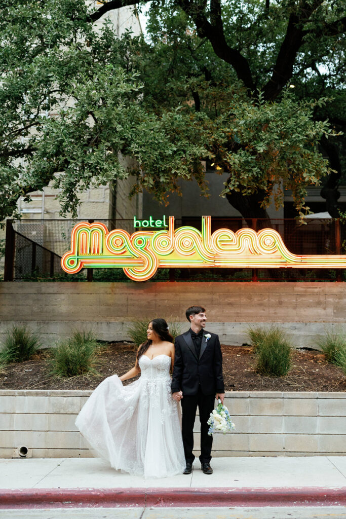 A bride and groom stand hand in hand in front of the neon "Hotel Magdalena" sign, with greenery and trees in the background. A wedding they planned from afar with their destination wedding planner, In Ink.
