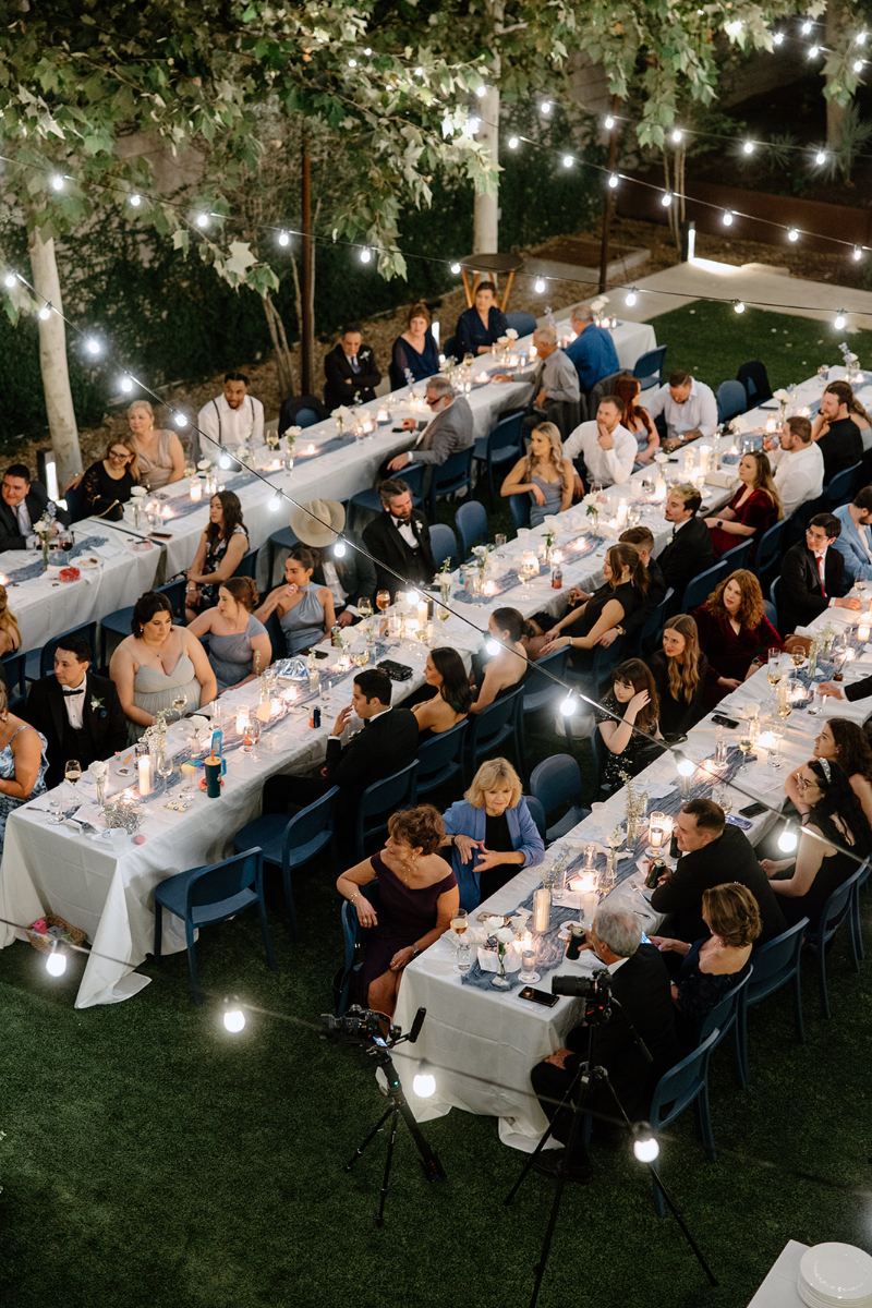 An overhead view of a formal outdoor reception planned by a destination wedding planner at night shows guests seated at long tables decorated with candles and string lights.