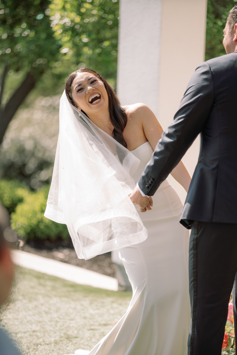 A bride in a white gown and veil laughs while holding hands with a man in a dark suit during an outdoor wedding ceremony.