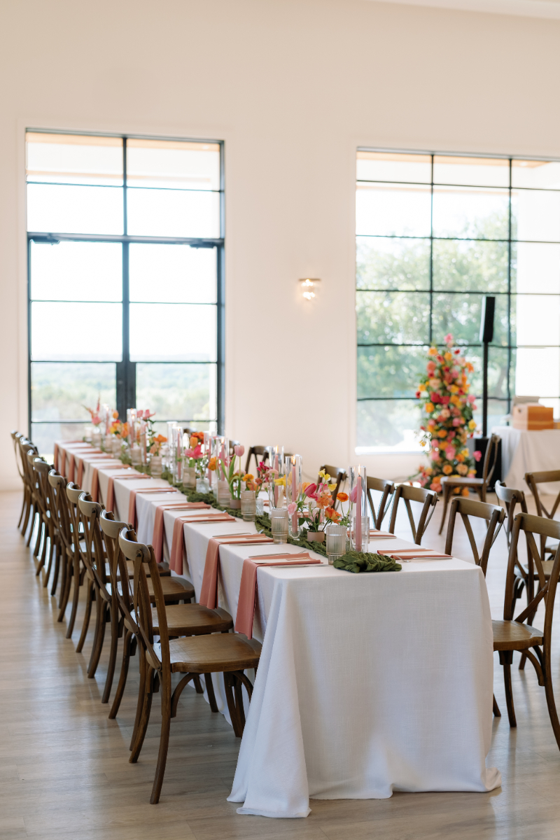 A long dining table with wooden chairs is set with pink and white decor, flowers, candles, and glassware in a bright room with large windows.