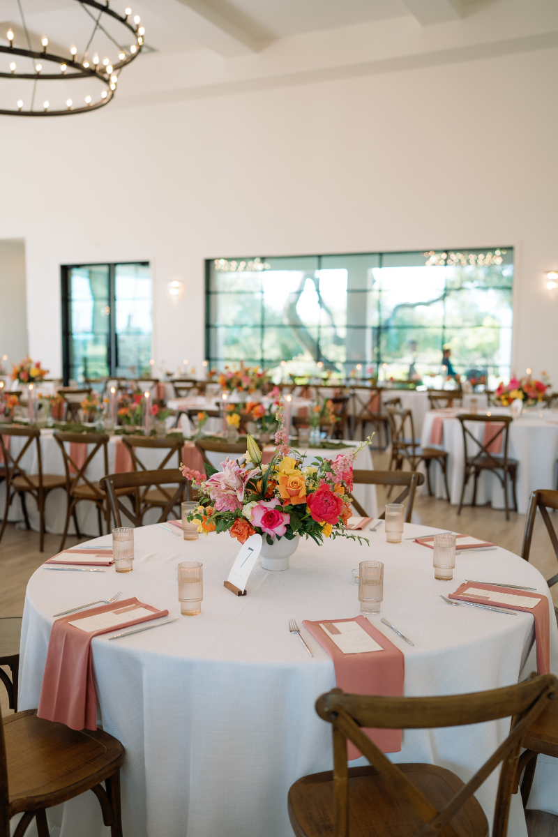 A round table with a floral centerpiece, pink napkins, and glassware is set in a bright event space with other similarly decorated tables in the background.