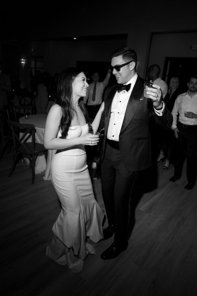 A woman in a strapless gown and a man in a tuxedo and sunglasses dance together at an indoor event, holding drinks and smiling.