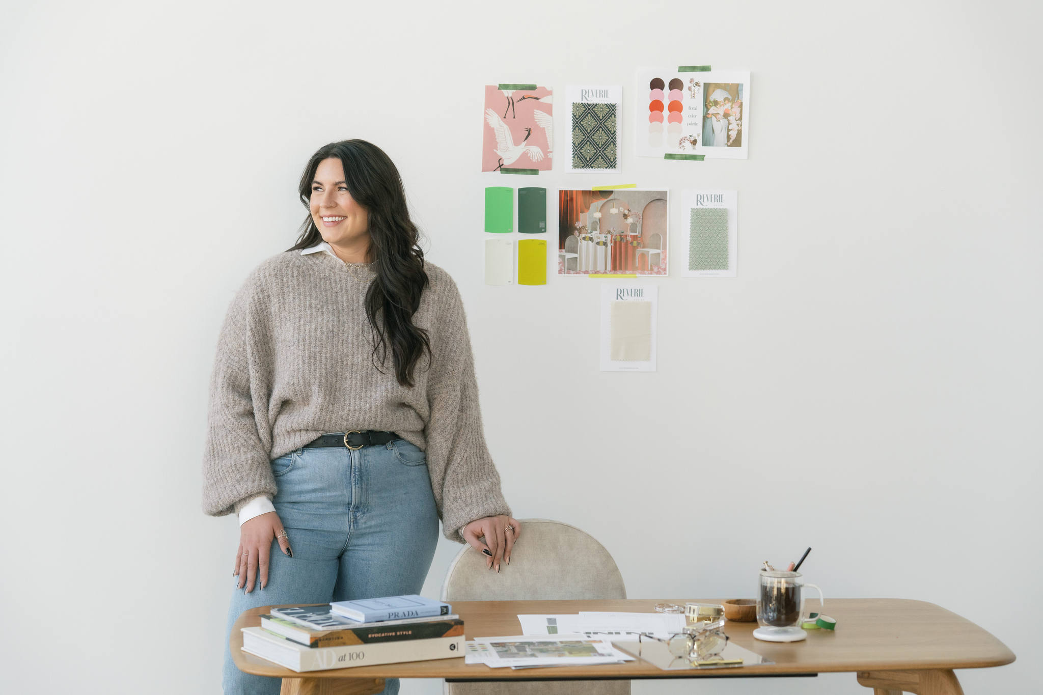Sydney of In Ink Weddings stands next to a desk with books and papers; design swatches and inspiration images are taped to the white wall behind her, talking about the wedding planning experience.
