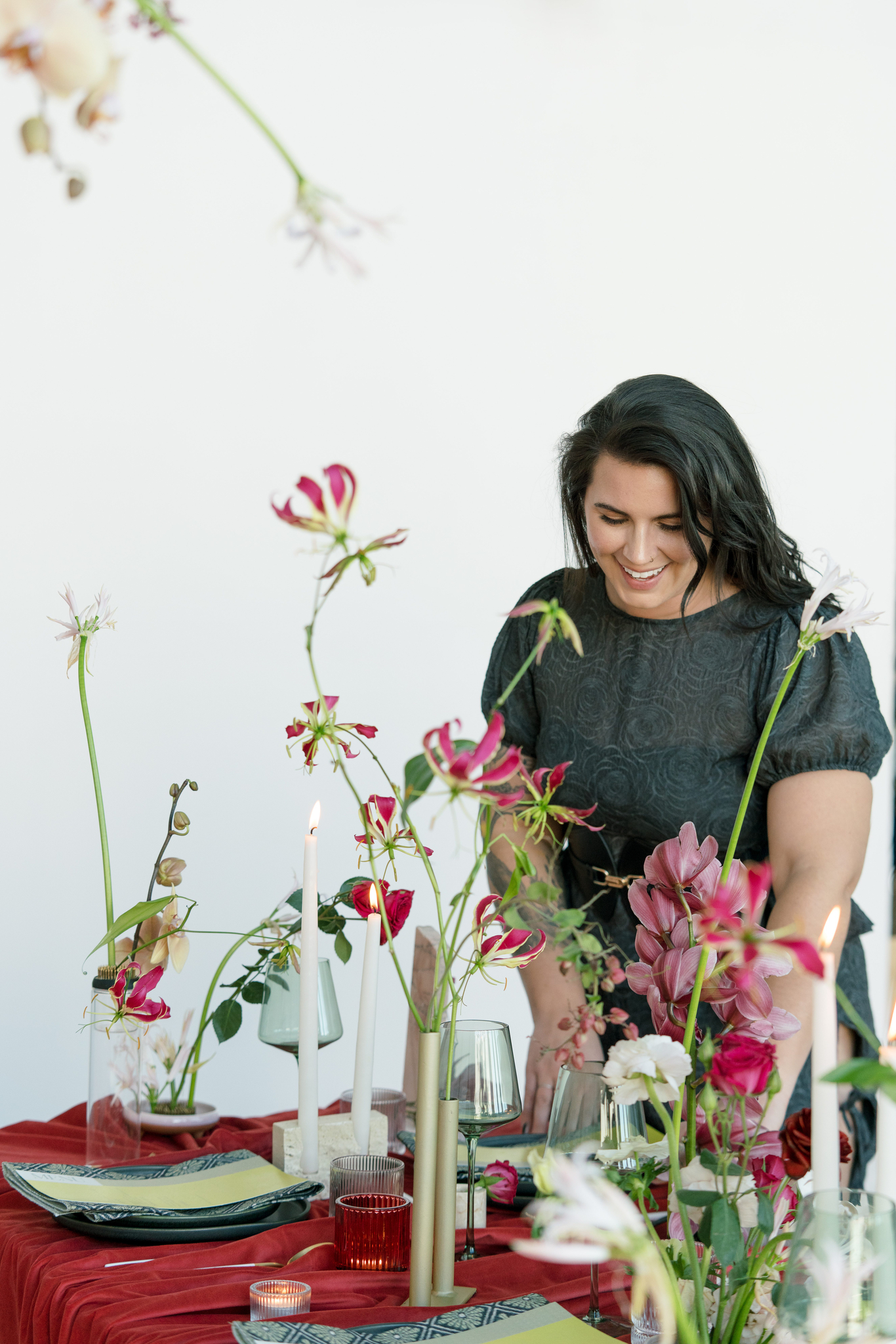 Sydney Meyer from In Ink Weddings, a full-service wedding planing business, in a dark dress arranges a table set with candles, plates, and various pink and white flowers on a red tablecloth.