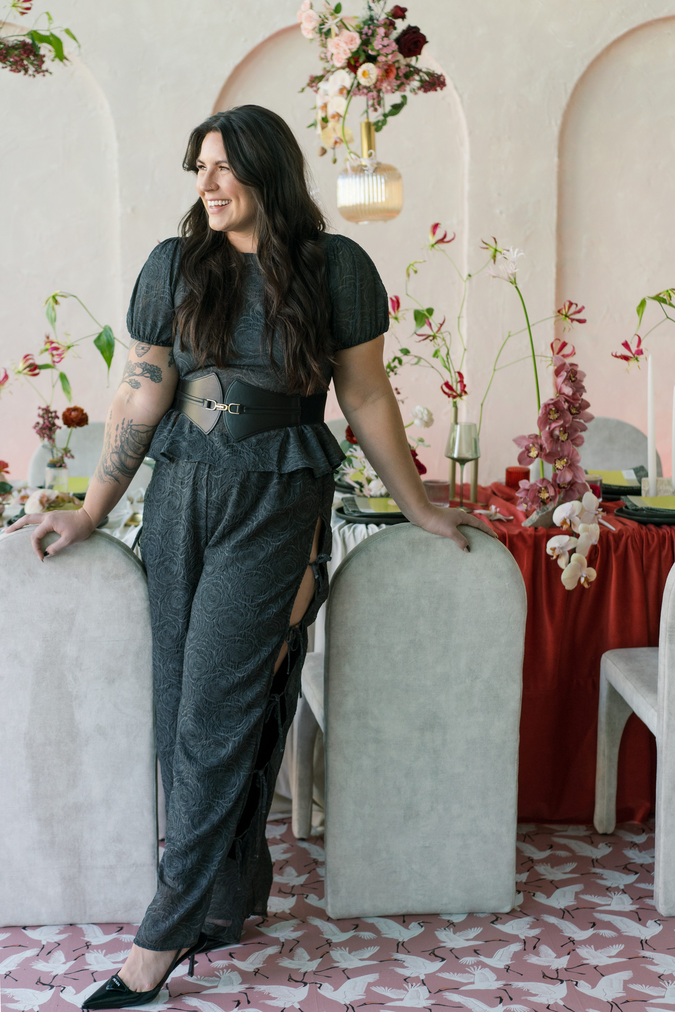 A woman with long dark hair stands smiling by a table decorated with flowers and candles; she wears a black patterned outfit and black heels.