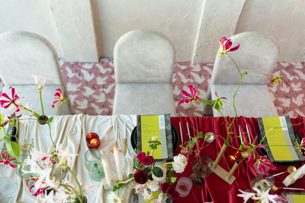 Overhead view of a table set with red and white tablecloths, floral arrangements, candles, and place settings, with three empty upholstered chairs.
