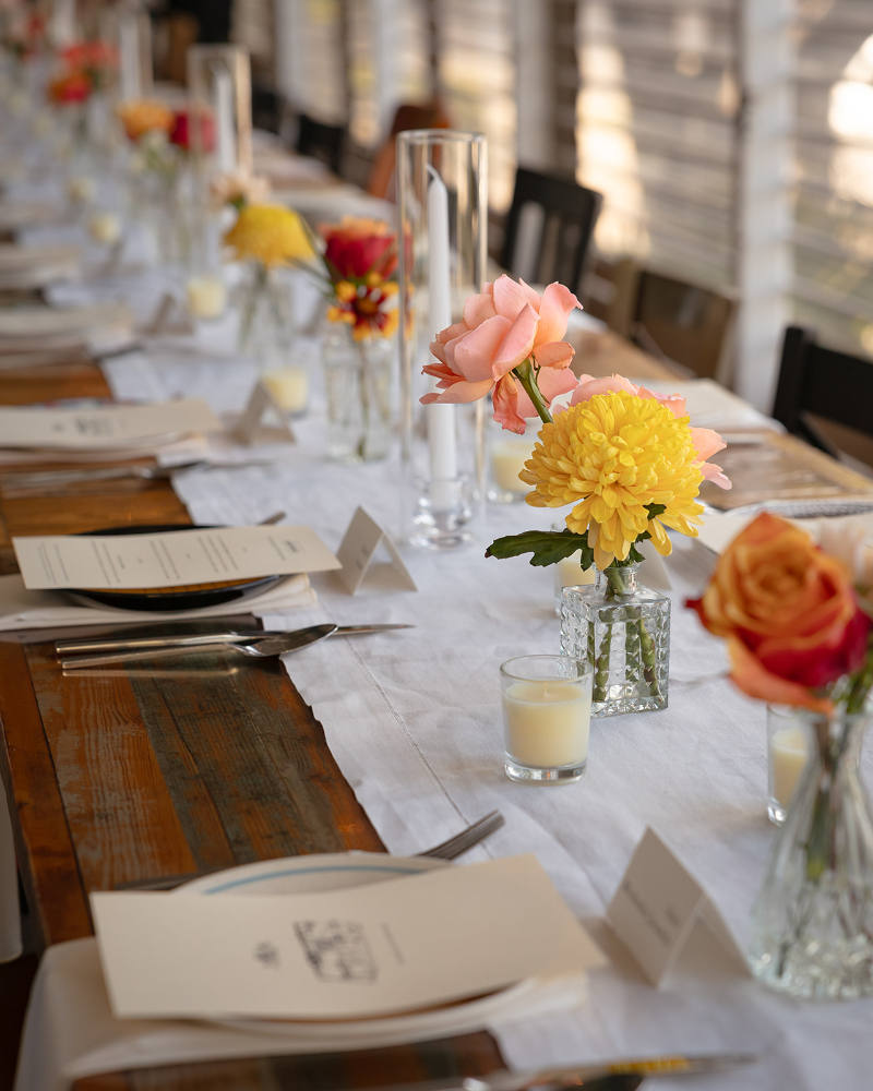 A long table set for a formal event, decorated with flowers in small vases, menus, name cards, and candles on a white table runner.