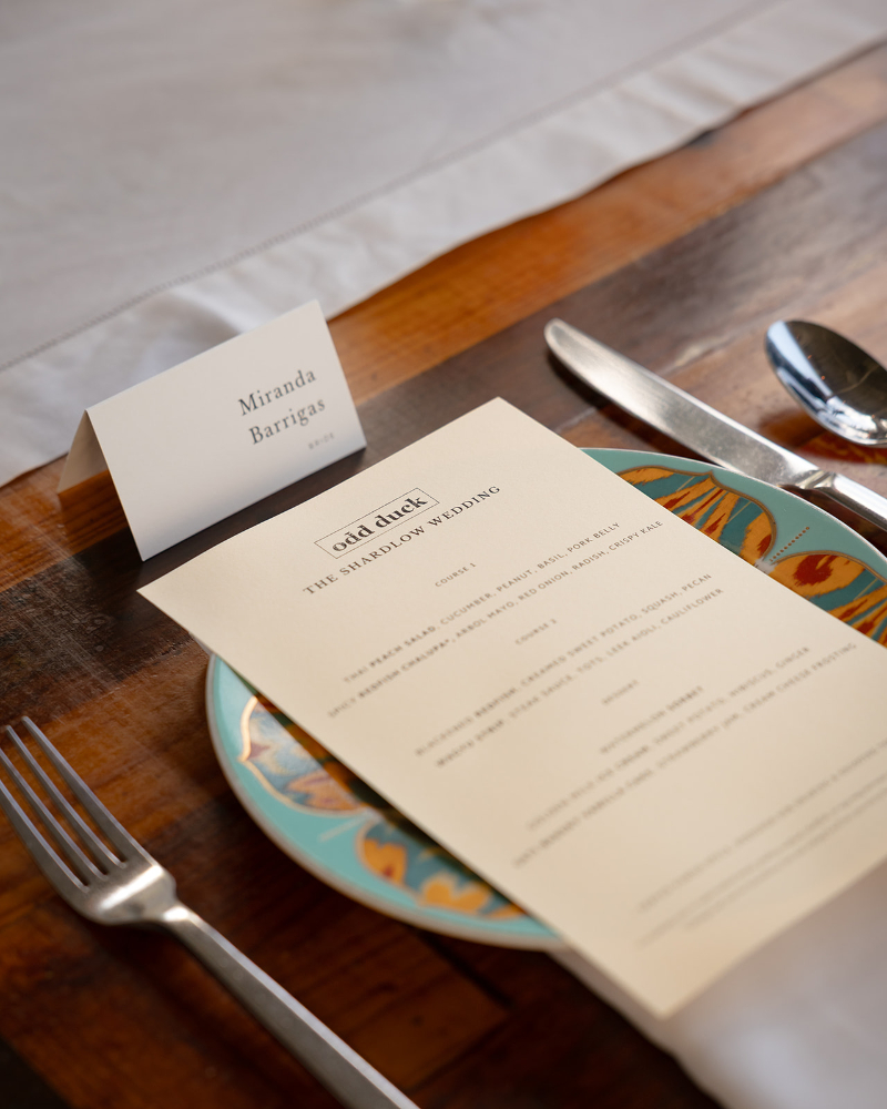 A wedding menu card and a name card reading "Miranda Barrigas" placed on a decorative plate, with cutlery set on a wooden table.