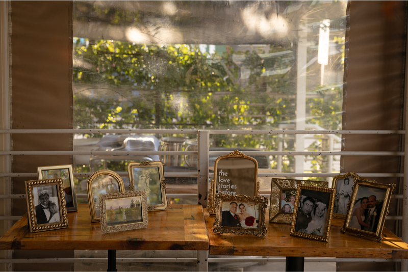 A collection of framed photographs is displayed on two wooden tables, set against a backdrop of trees and sunlight streaming through a clear barrier.