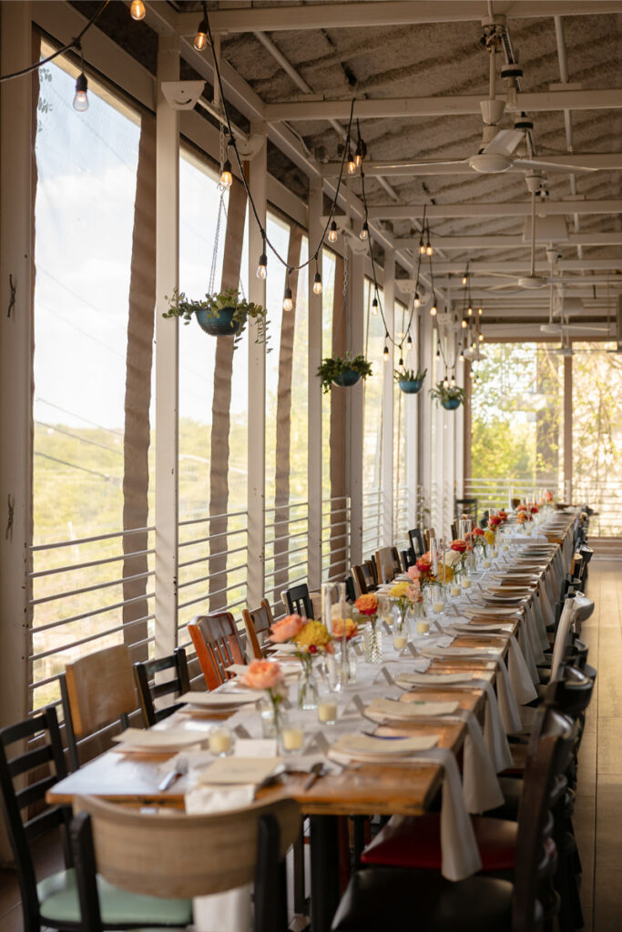 A long dining table is set with plates, glasses, and floral centerpieces in a sunlit room with hanging plants and string lights.