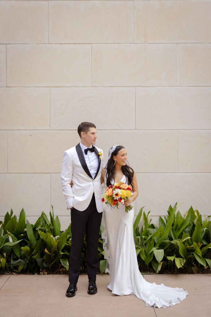 A bride in a white dress holding a bouquet stands beside a groom in a white tuxedo jacket and black pants, both facing slightly left, with green plants and a beige stone wall behind them.