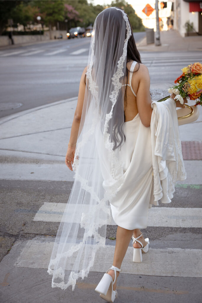 A woman in a white dress and veil walks across a street, holding a bouquet of flowers and a draped garment.