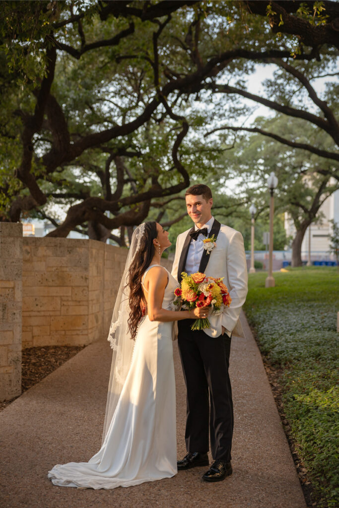 A bride in a white dress and veil stands beside a groom in a white tuxedo jacket, holding a bouquet, outdoors under large trees.