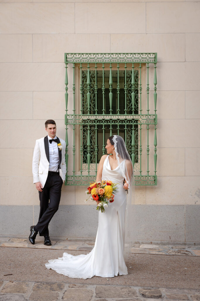 A bride in a white dress and veil holding a bouquet stands near a groom in a white jacket and black pants, both posing in front of a beige wall with a green, ornate window grill.