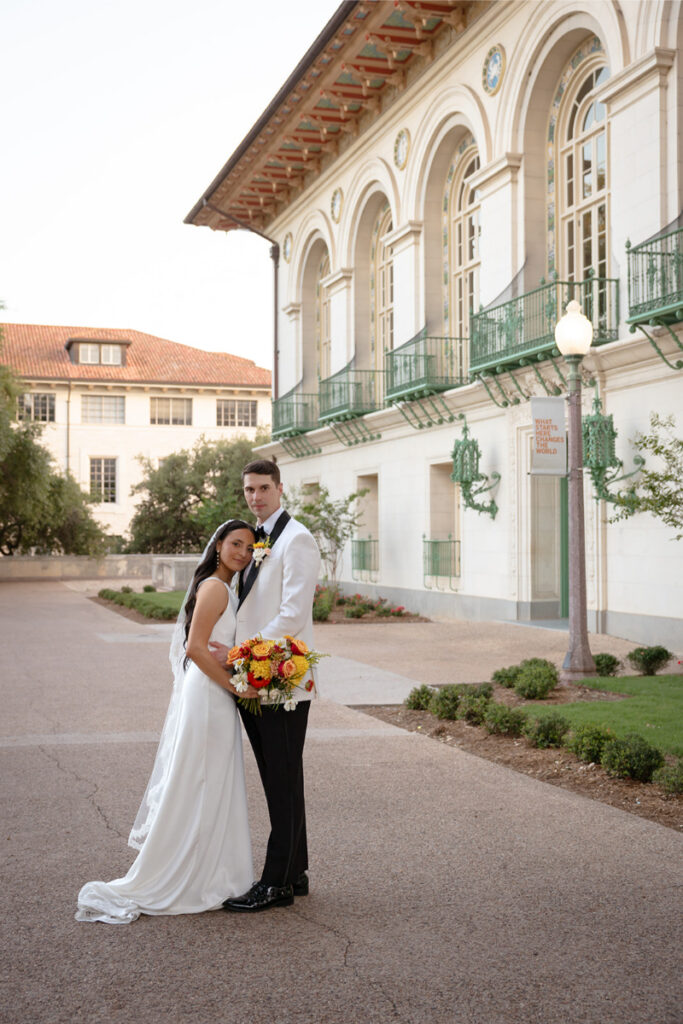 A bride in a white dress and groom in a white tuxedo jacket stand together outside a historic building after their Santa Barbara courthouse wedding, holding a bouquet of flowers.