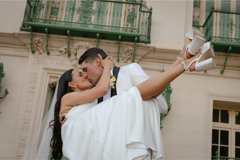 A groom in a white suit lifts and kisses a bride in a white dress and high heels after their Santa Barbara courthouse wedding.