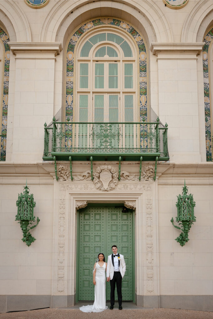 A bride and groom stand side by side in front of a large green door with ornate architectural details and decorative mosaic panels above after their Santa Barbara courthouse wedding.