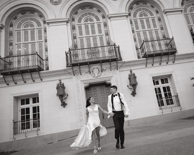 A bride and groom in formal attire hold hands and walk outside their Santa Barbara courthouse wedding with tall arched windows and ornate balconies.