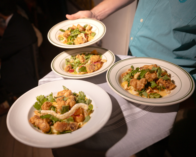 A person wearing a teal shirt holds four plates of fried chicken salad garnished with greens and herbs on a tray covered with a white cloth.