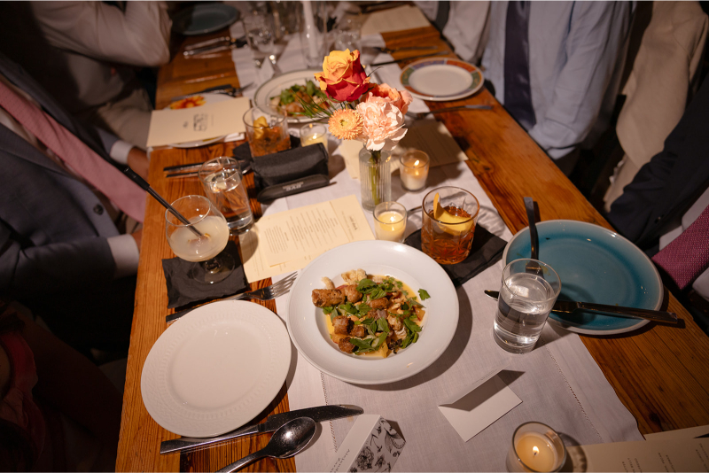 A wooden table set for a meal with salads, drinks, plates, candles, flowers in a vase, and people seated nearby, some dressed in suits.