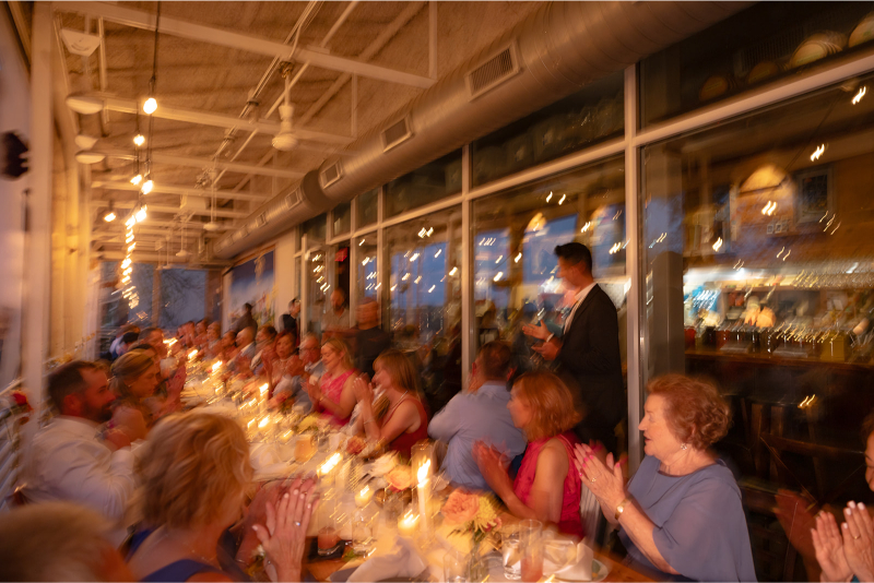 A large group of people seated at a long, candlelit table indoors, applauding a person standing and speaking near the window; the image is blurred.