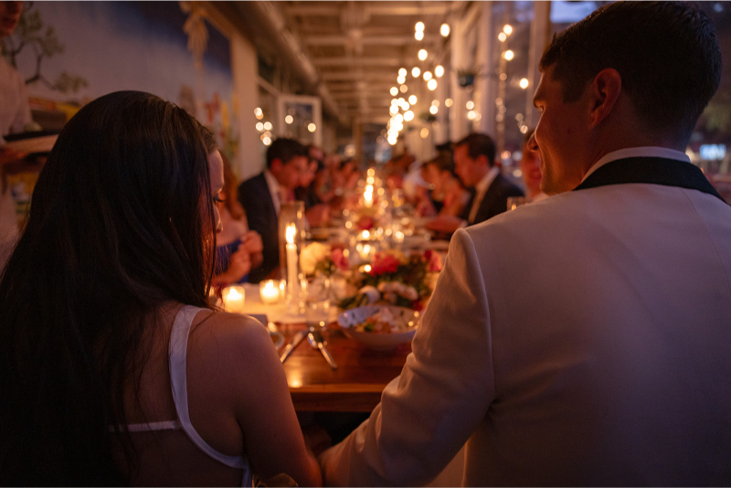 A couple sits at the head of a long, candlelit dinner table surrounded by guests in formal attire at an evening event.