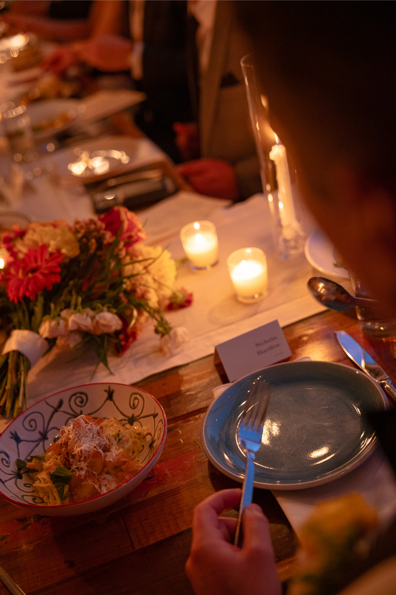 A person sits at a wooden table set for a formal dinner with candles, flowers, a bowl of pasta, a blue plate, and a place card visible.