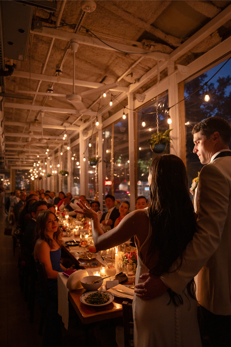 A bride and groom stand at the head of a long dinner table, addressing seated guests at an evening indoor wedding reception with string lights and large windows.