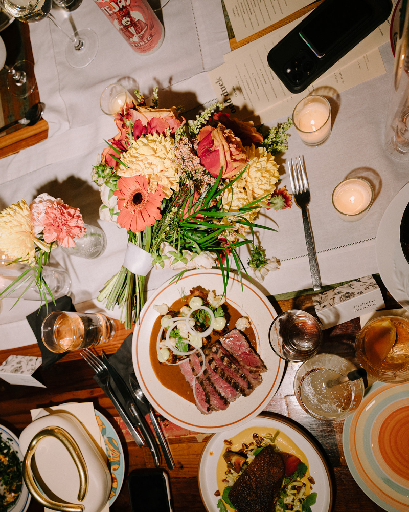 A dining table set with plates of food, a bouquet of flowers, drinks, menus, candles, and a phone arranged on a white table runner.