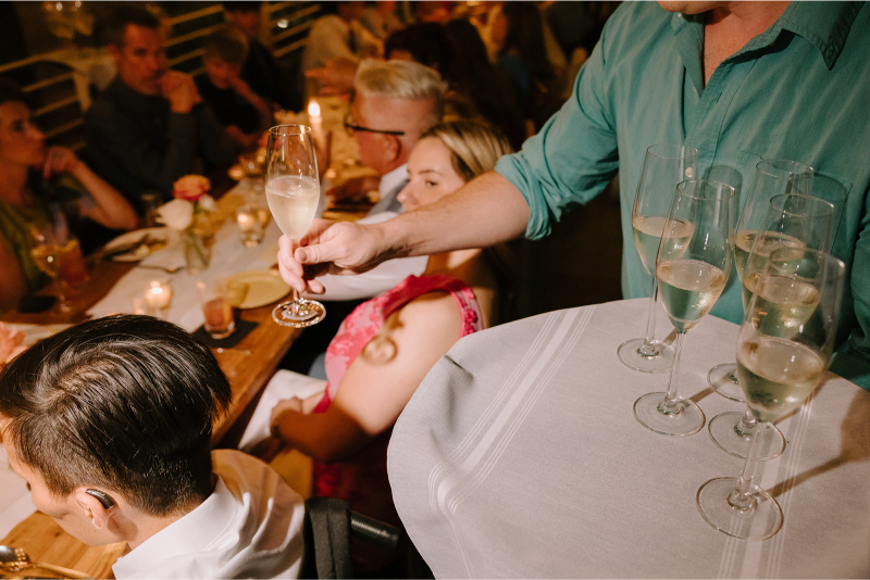 A server in a teal shirt hands a glass of champagne to a seated guest at a crowded, candle-lit dinner event.