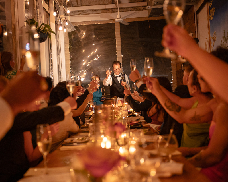 A group of people seated at a long table indoors raise glasses in a toast, with a person standing at the head of the table leading the toast.