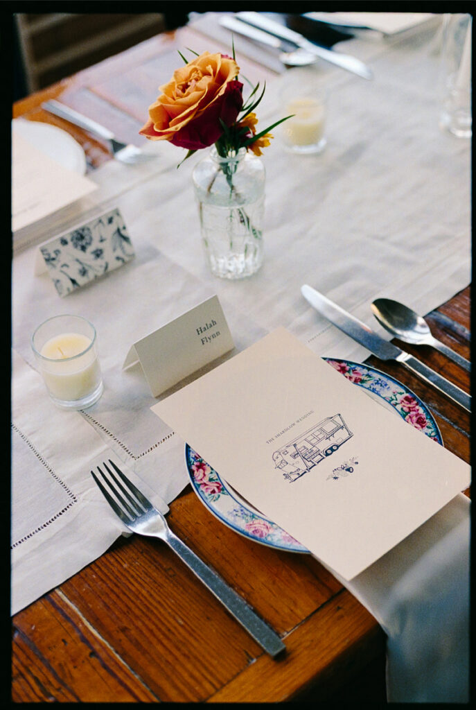 A table setting with a floral plate, menu card, name card, silverware, a small glass vase with orange roses, and a votive candle on a wooden table.