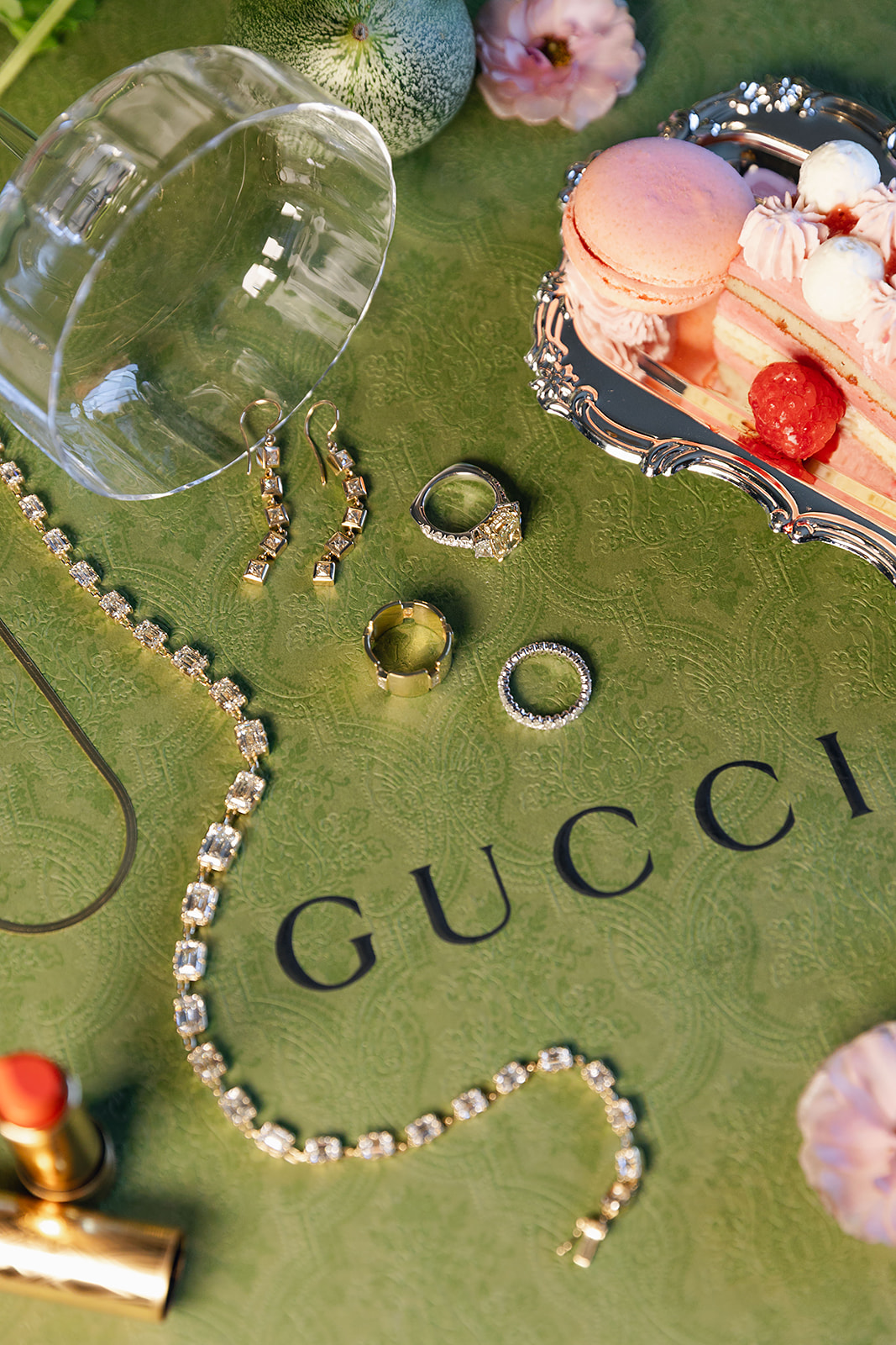 Jewelry, earrings, rings, a diamond necklace, and a gold tube of lipstick are displayed on a green Gucci surface alongside a tipped glass and a pastry on a silver tray.