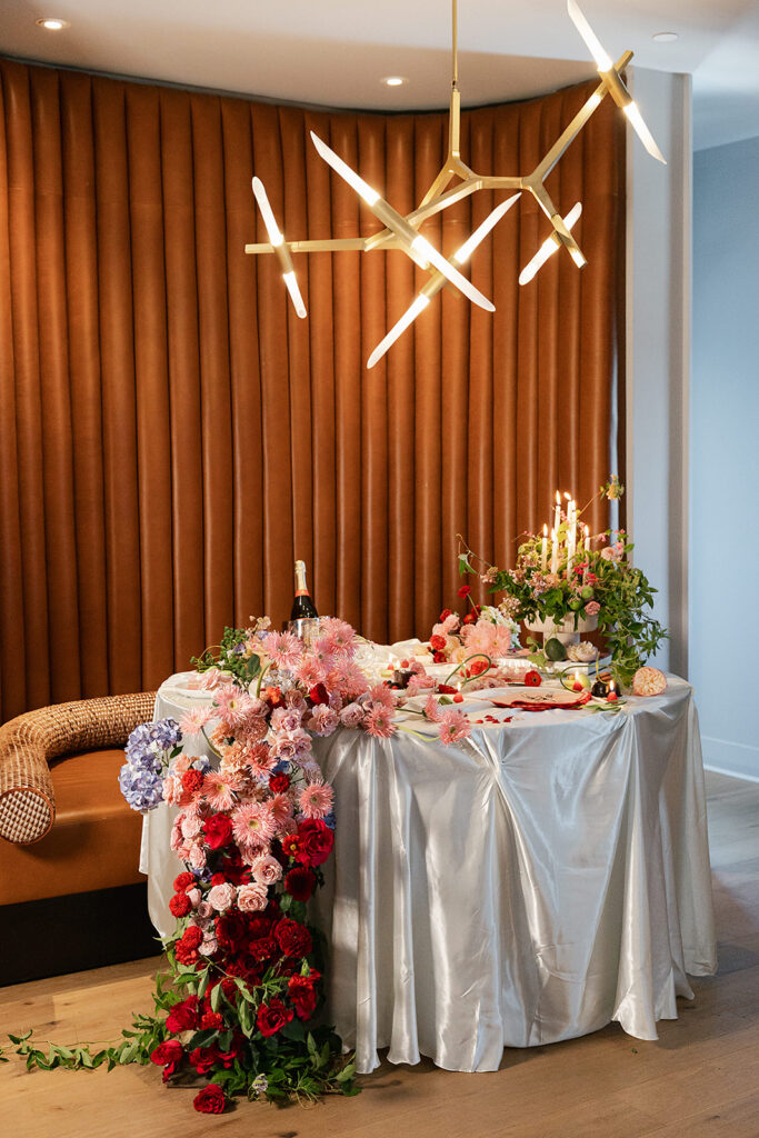 A round table with a white satin tablecloth decorated with assorted flowers, candles, and a bottle of champagne, set beneath a modern gold chandelier, all assembled with a full service wedding planning team.