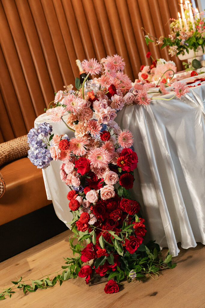 A white table with a cascading floral arrangement of pink, red, and purple flowers, set against a brown upholstered wall and wooden floor, all assembled with a full service wedding planning team.