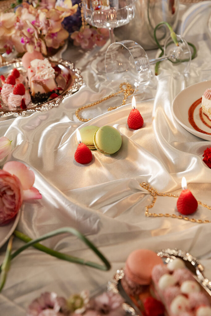 Two green macarons, raspberries, and small candles are arranged on a silky white tablecloth, surrounded by desserts, flowers, and an overturned glass.