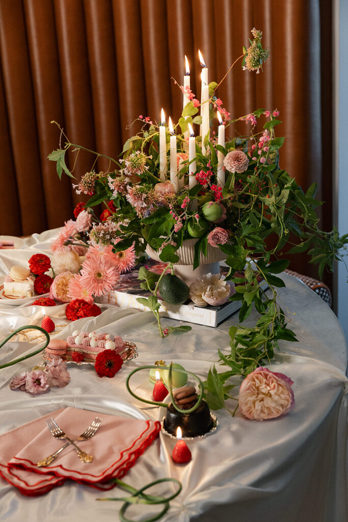 A round table set with pink flowers, greenery, red strawberries, lit white candles, pink napkins, and desserts on a white tablecloth in front of a brown curtain.