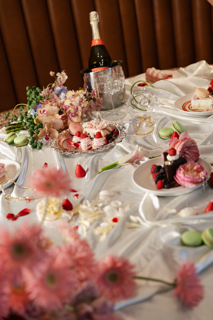 A table with desserts, macarons, strawberries, flowers, a bottle of sparkling wine, two glasses, and jewelry, all arranged on a white satin tablecloth.