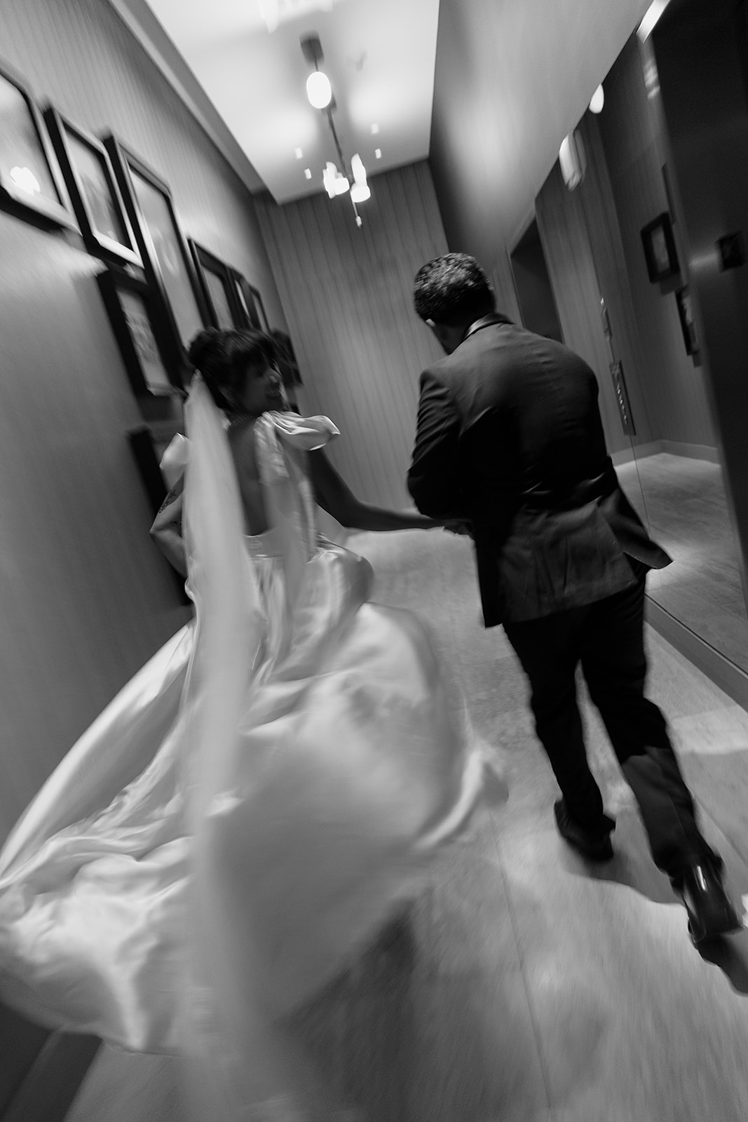 A bride and groom, seen from behind, run down a hotel hallway. The image is in black and white and slightly blurred, showing movement.