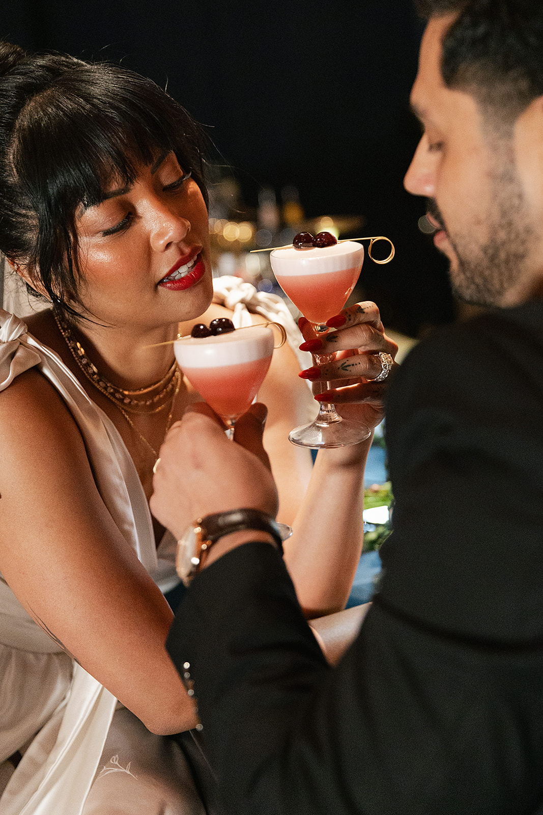 A bride in a white dress and a groom in a dark suit clink glasses of pink cocktails garnished with cherries, they are in the moment thanks to full service wedding planning.