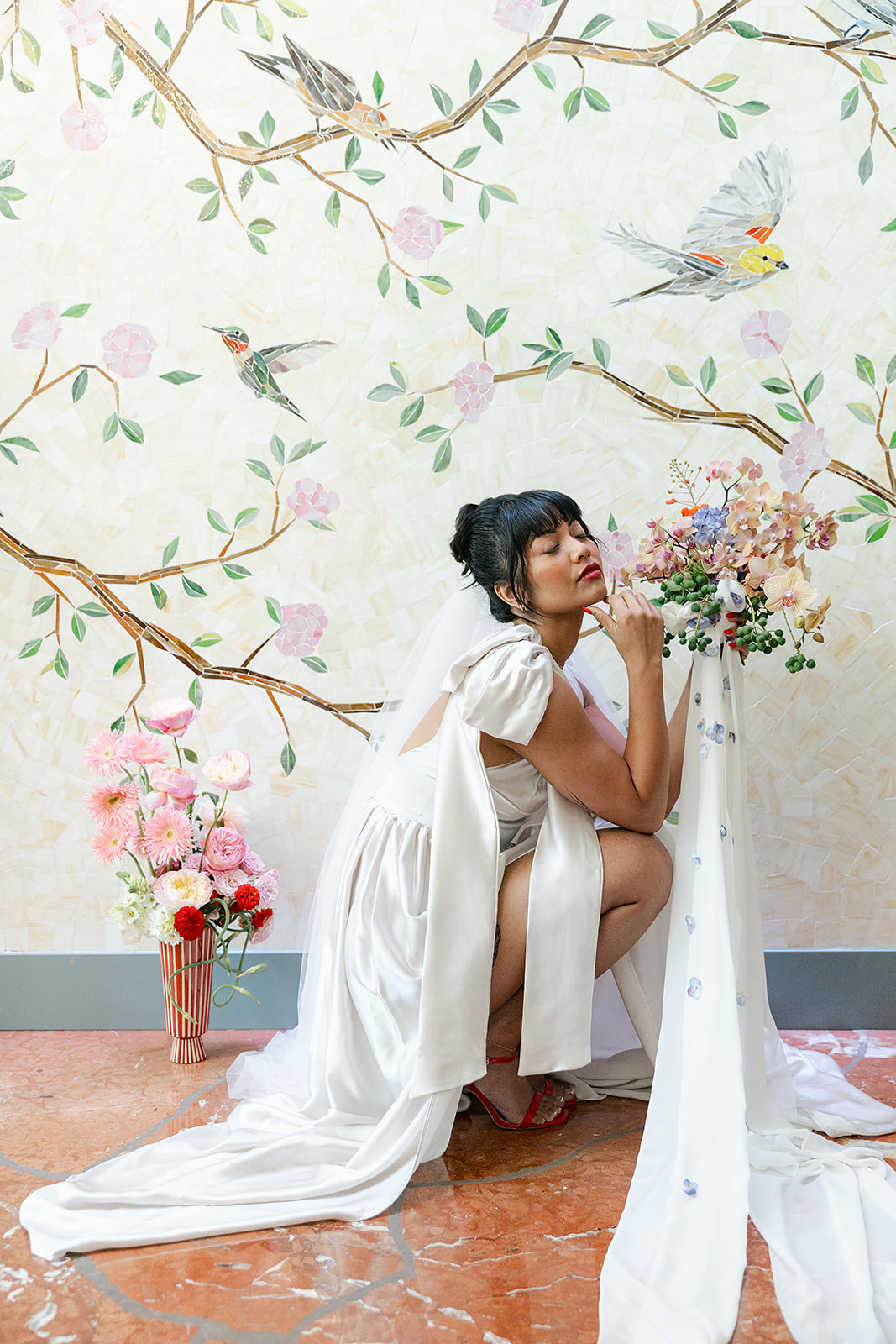 A woman in a white dress and veil kneels next to a floral arrangement, with a painted wall of birds and branches in the background.