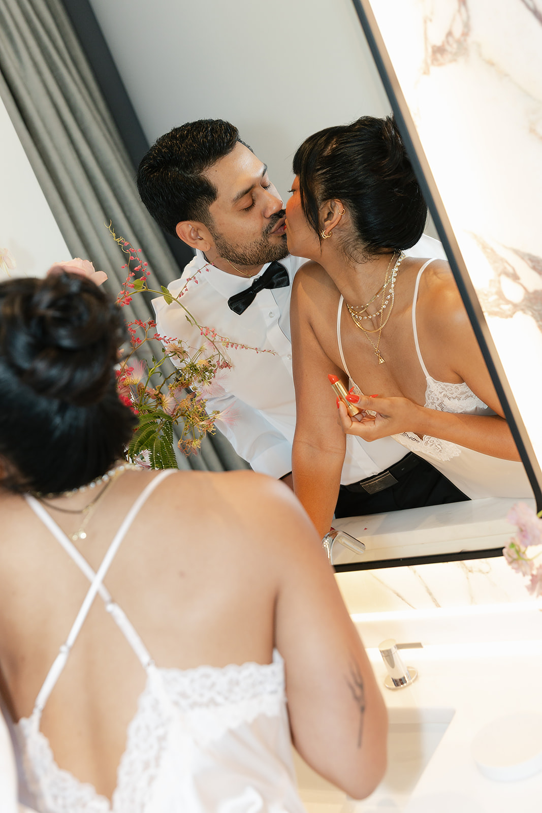 A woman in a white lace camisole applies lipstick at a bathroom sink while sharing a kiss with a man in a white shirt and bow tie. Both are reflected in the mirror.
