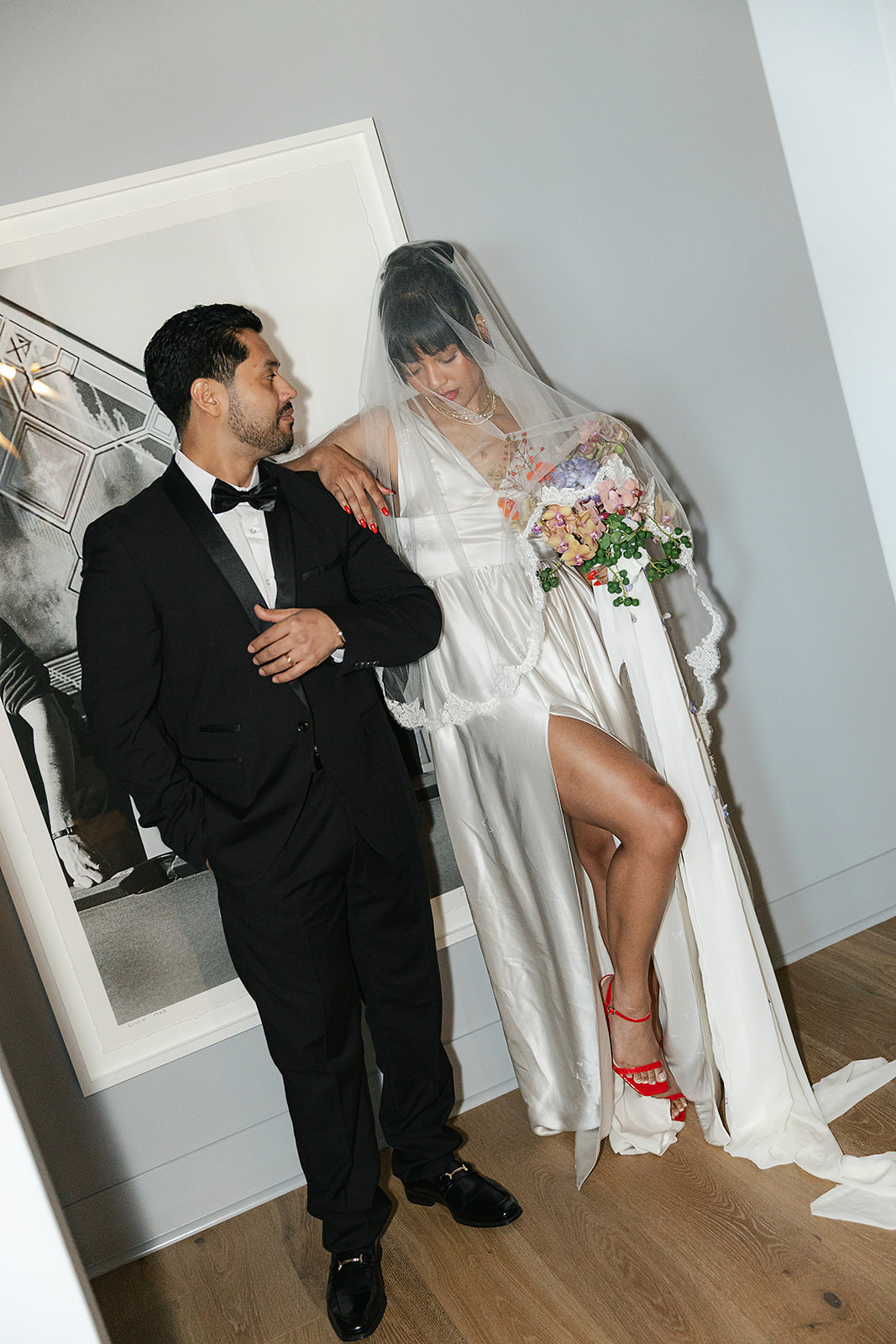 A bride in a white dress and veil stands beside a groom in a black tuxedo; she holds a bouquet and lifts her dress to show red heels.