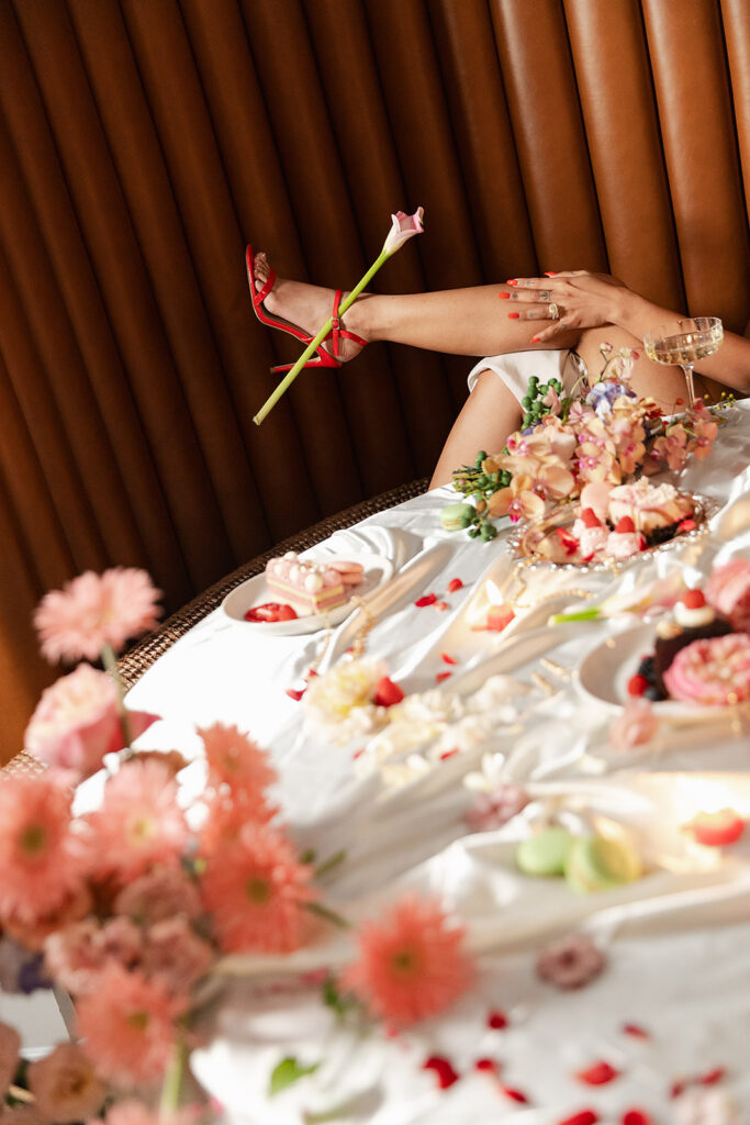 A person with crossed legs in red heels holds a pink flower, reclining behind a table set with flowers, desserts, and drinks on a white tablecloth.