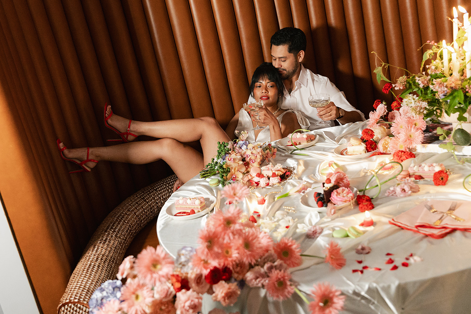 A couple lounges on a curved bench beside a table covered with flowers, candles, and food. The woman leans back with her legs up as they both hold glasses, surrounded by a decorative arrangement put together with full service wedding planning.