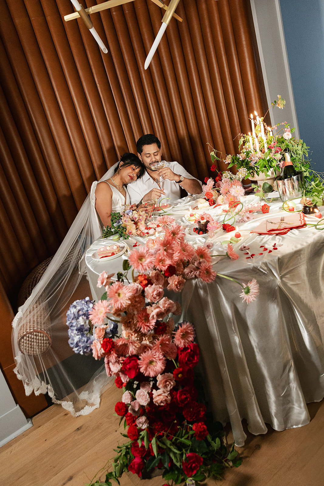 A bride and groom sit closely at a decorated table with flowers, candles, and scattered petals, appearing relaxed and happy after a wedding celebration that was made possible with full service wedding planning.
