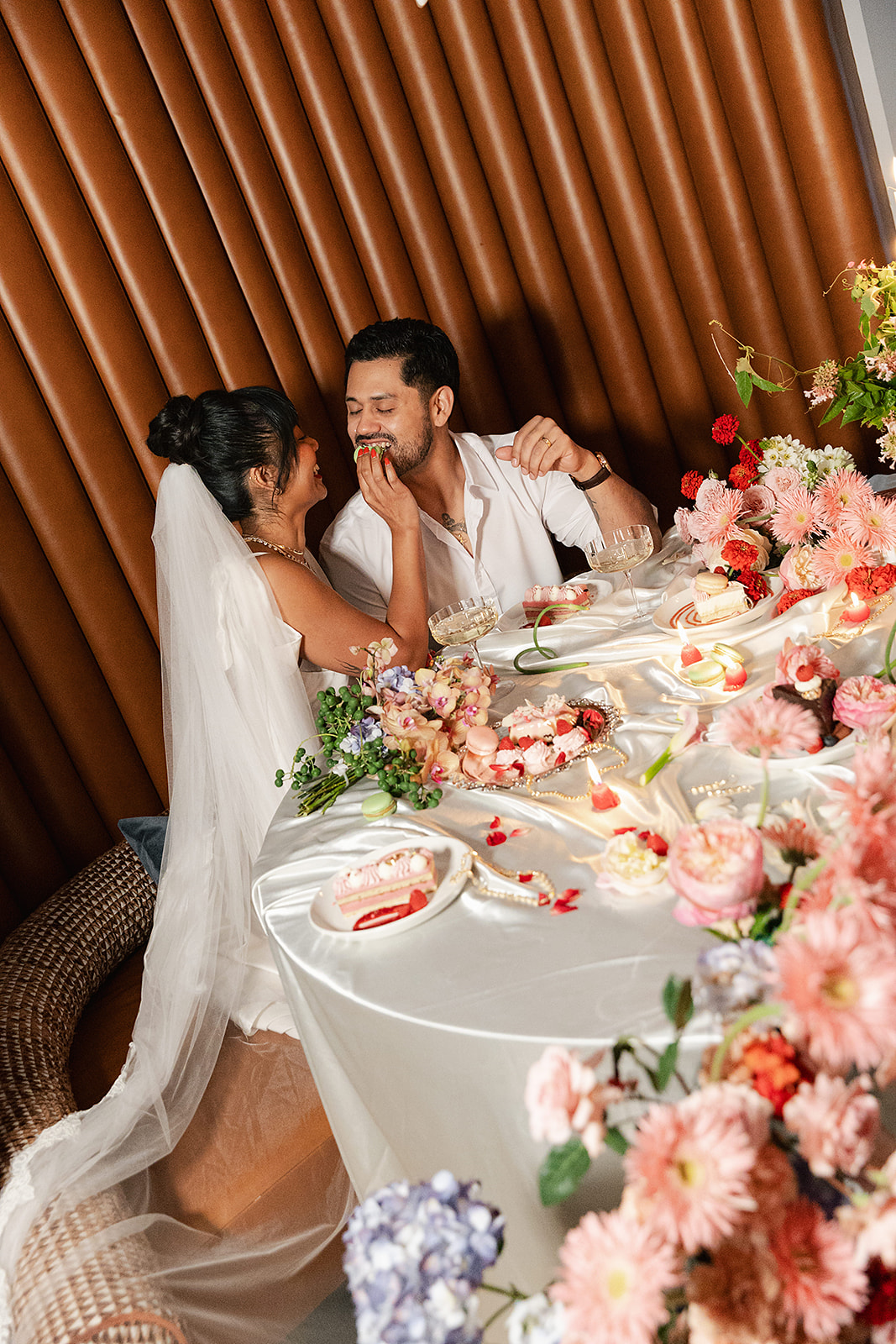 A bride feeds cake to a groom at a decorated table with flowers and desserts, both smiling and enjoying the moment that was made possible with full service wedding planning.