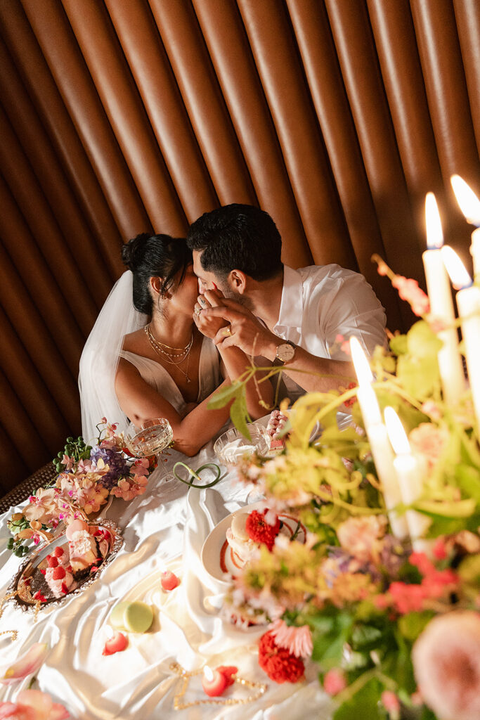 A bride and groom share a kiss at a decorated table set with candles, flowers, and food, with the bride wearing a veil and white dress, all made possible with a full service wedding planning team.