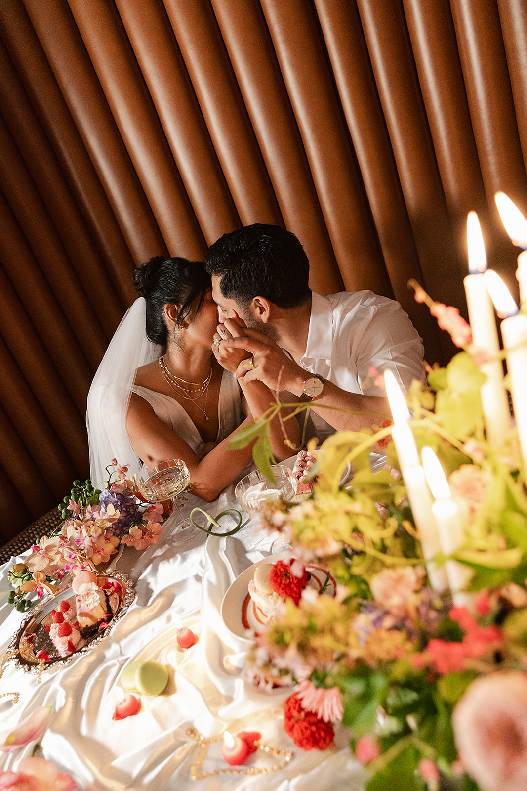 A bride and groom share a kiss at a decorated table set with candles, flowers, and food, with the bride wearing a veil and white dress.