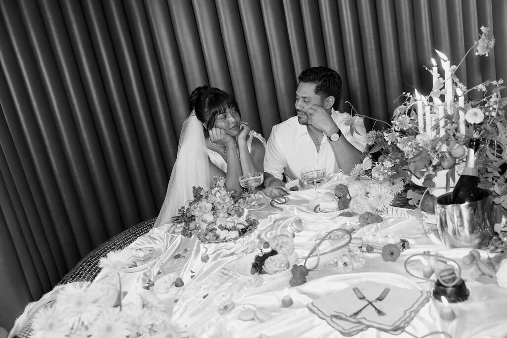 A bride and groom sit at a decorated table, looking at each other with relaxed expressions, surrounded by food, flowers, and candles, assembled with a full service wedding planning team.
